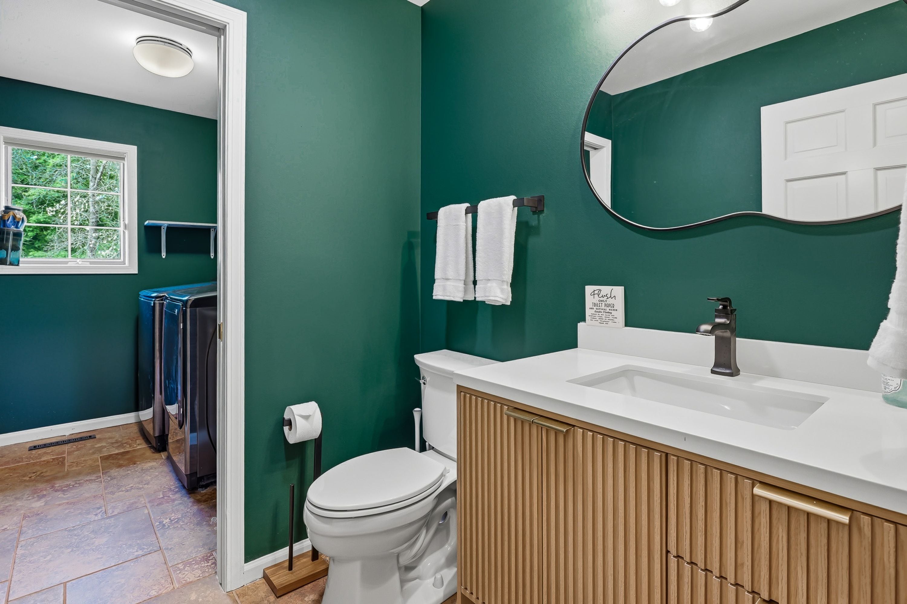 A bathroom with a white sink, a toilet, and a mirror. There is a window with a view of trees and a shelf with bottles.