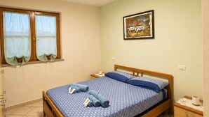 A single-bed room with a wooden frame, a patterned bedspread, a small bedside table with a lamp, a window with curtains, and a framed picture on the wall. - Sunset in Seu, 150 meters from the beach. Ideal for families and couples. (Funtana Meiga)
