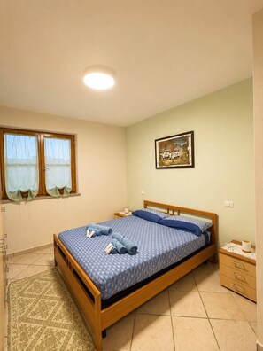 Ein Einzelzimmer mit einem Holzbett, einer gemusterten Tagesdecke, einem kleinen Nachttisch mit Lampe, einem Fenster mit Vorhängen und einem gerahmten Bild an der Wand.
