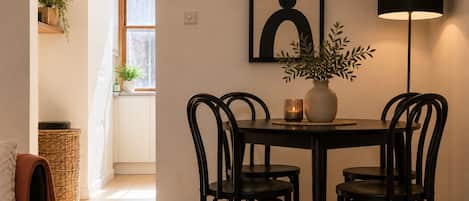 A dining area with a round table, four chairs, a vase with plants, a framed artwork, and a floor lamp.