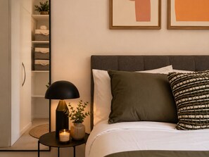 Ein Schlafzimmer mit einem Bett, einem Nachttisch mit Lampe, einer Vase mit Pflanze und einem Regal mit gefalteten Handtüchern.