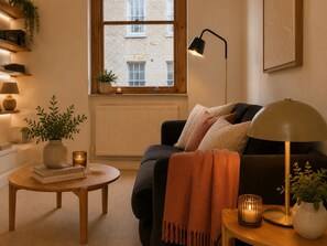 Een gezellige woonkamer met een bank, een klein rond tafeltje, een vloerlamp en een raam met uitzicht op een gebouw.