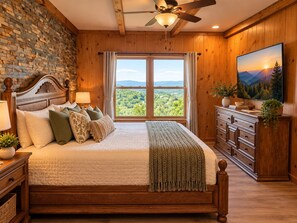 Ein Schlafzimmer mit einem großen Bett, einem Holzkopfende, einem Deckenventilator, Blick ins Freie durch ein Fenster und einem Flachbildfernseher an der Wand.