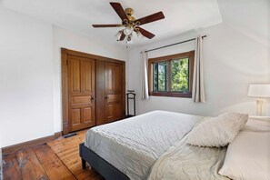 Ein Schlafzimmer mit Bett, Deckenventilator, Fenster mit Vorhängen und einer Holztür.