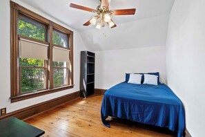 A bedroom with a bed, a ceiling fan, a window with blinds, and a bookshelf. - The Blue Victorian | Near Downtown Traverse City (Traverse City)