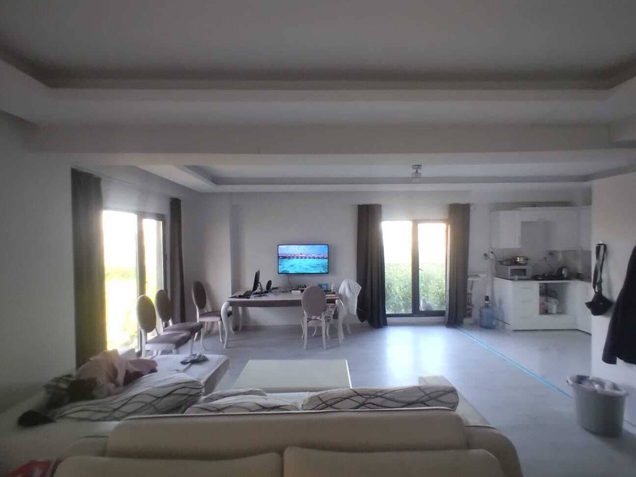 A spacious living area with a sofa, a dining table, chairs, a TV, and a kitchen in the background.