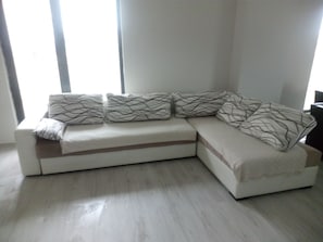 Un canapé d’angle avec des coussins à motifs, un plancher de couleur claire et une fenêtre avec des stores.