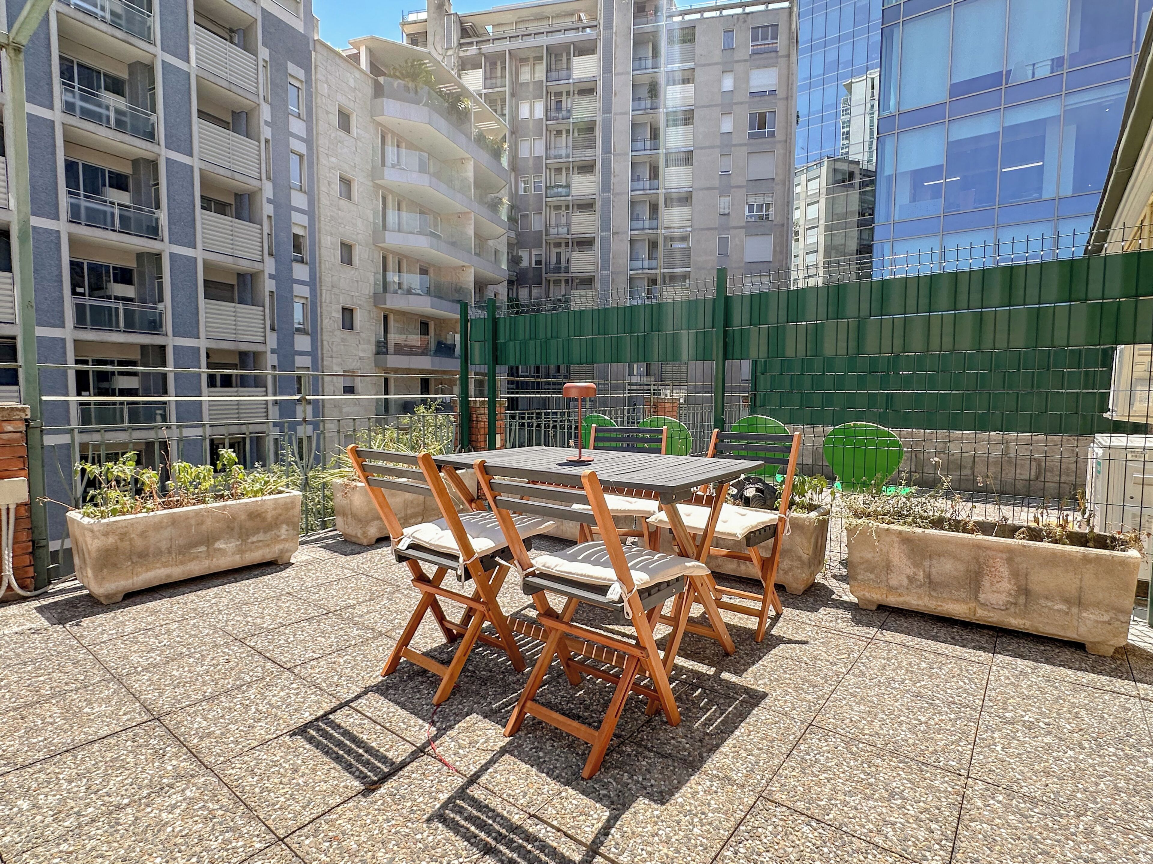 An outdoor patio with a table and chairs, surrounded by planters and a green fence.