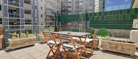 An outdoor patio with a table and chairs, surrounded by planters and a green fence.