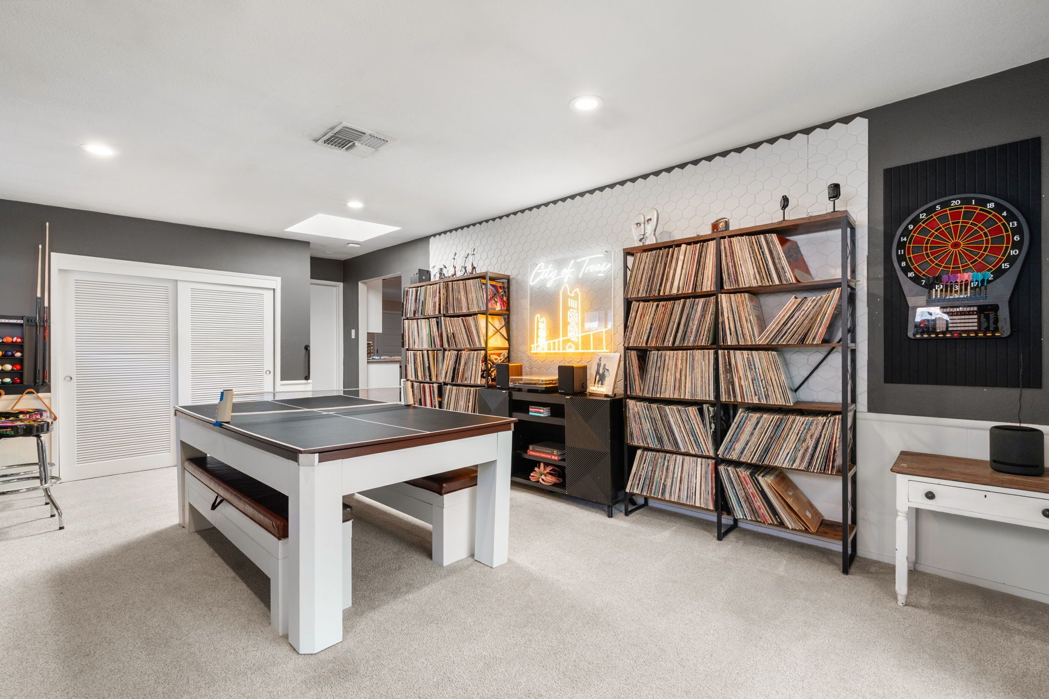 A room with a pool table, bookshelves, a dartboard, and a neon sign.