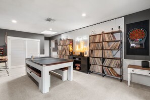 A room with a pool table, bookshelves, a dartboard, and a neon sign.