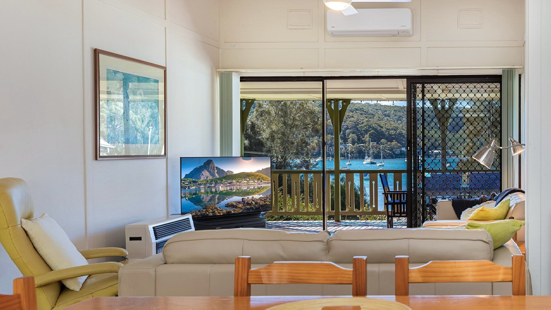 A living room with a sofa, a TV, and a table. There is a balcony with a view of the sea and boats.