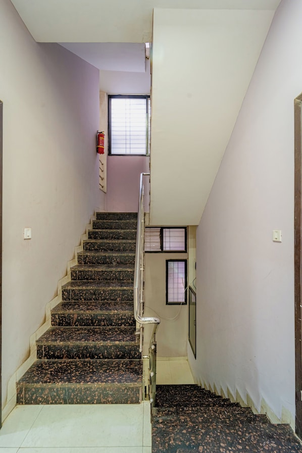 A staircase with a handrail, a fire extinguisher mounted on the wall, and a window with blinds. - Hotel Evanka by Navvarsh (Indore)