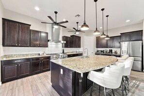 A modern kitchen with dark wood cabinets, granite countertops, a central island with bar stools, a stainless steel refrigerator, and ceiling fans. - Modern 5BR Pool Home Sleeps 15 Walk to Stadium (Glendale)