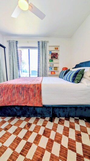 Ein Schlafzimmer mit Bett, Deckenventilator, Fenster mit Vorhängen, einem karierten Boden und einem Regal mit Büchern.