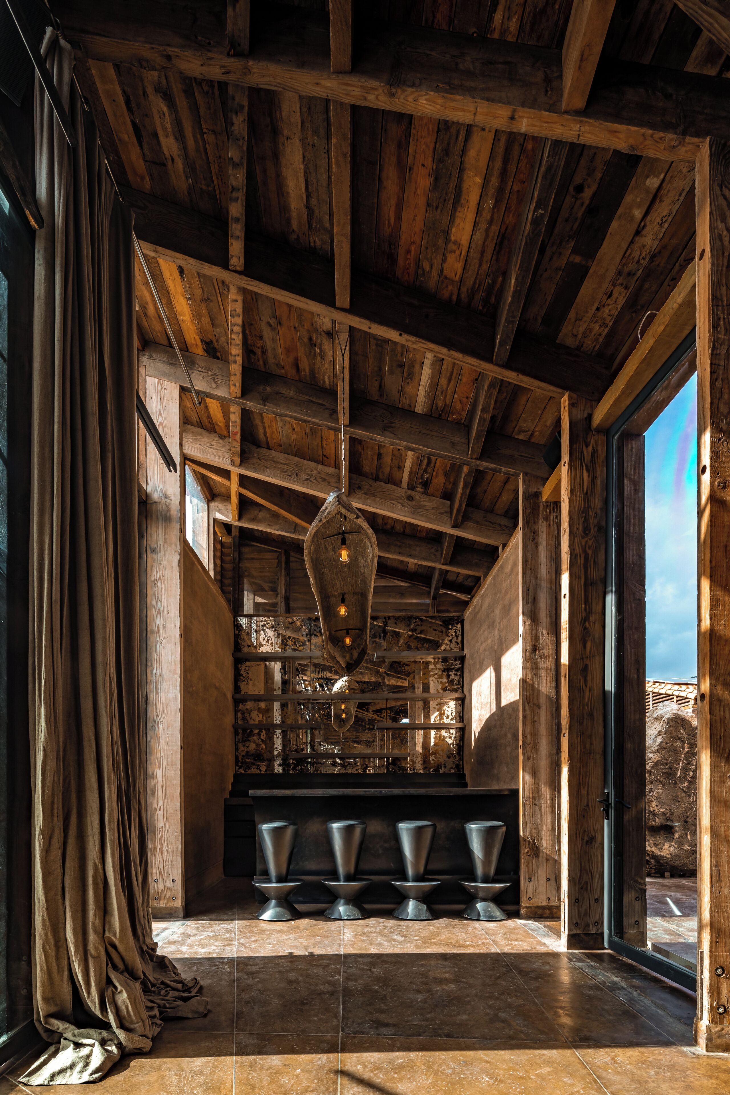 Un interior rústico con vigas de madera a la vista, una lámpara colgante y una zona de bar con taburetes.