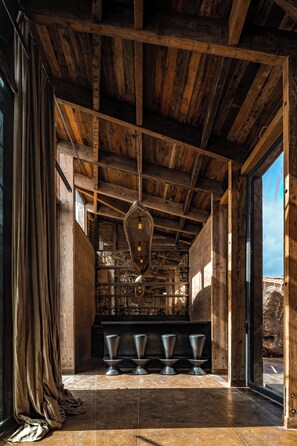 Um interior rústico com vigas de madeira aparente, um lustre pendurado e um espaço de bar com banquetas.
