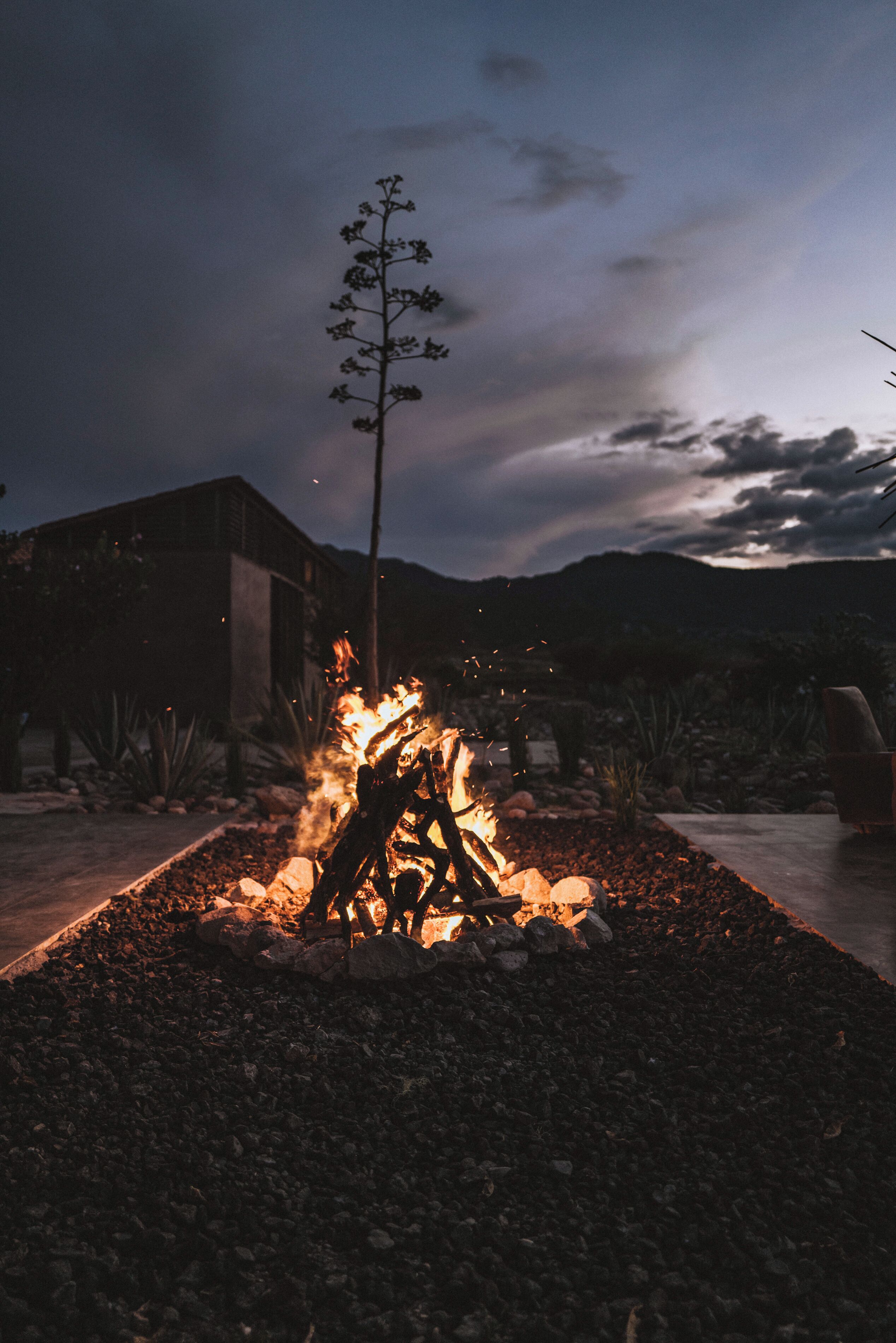A campfire with rocks around it, a building in the background, and a tree.