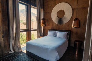 Un dormitorio con cama, un ventanal amplio, cortinas, una lámpara de pared y vistas a un paisaje montañoso.