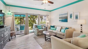 A spacious living room with a ceiling fan, a sofa set, a coffee table, and a television. There is a sliding glass door leading to a balcony with outdoor furniture. - 3BR Ocean Breeze Escape | Walk to Beach at Coligny (Hilton Head Island)