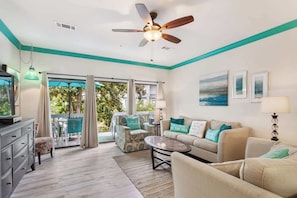 A spacious living room with a ceiling fan, a sofa set, a coffee table, and a television. There is a sliding glass door leading to a balcony with outdoor furniture. - 3BR Ocean Breeze Escape | Walk to Beach at Coligny (Hilton Head Island)