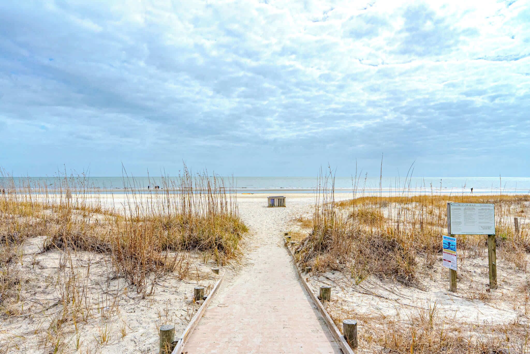 Ein Sandweg am Strand, der zum Meer führt, mit einer Bank und Informationstafeln.