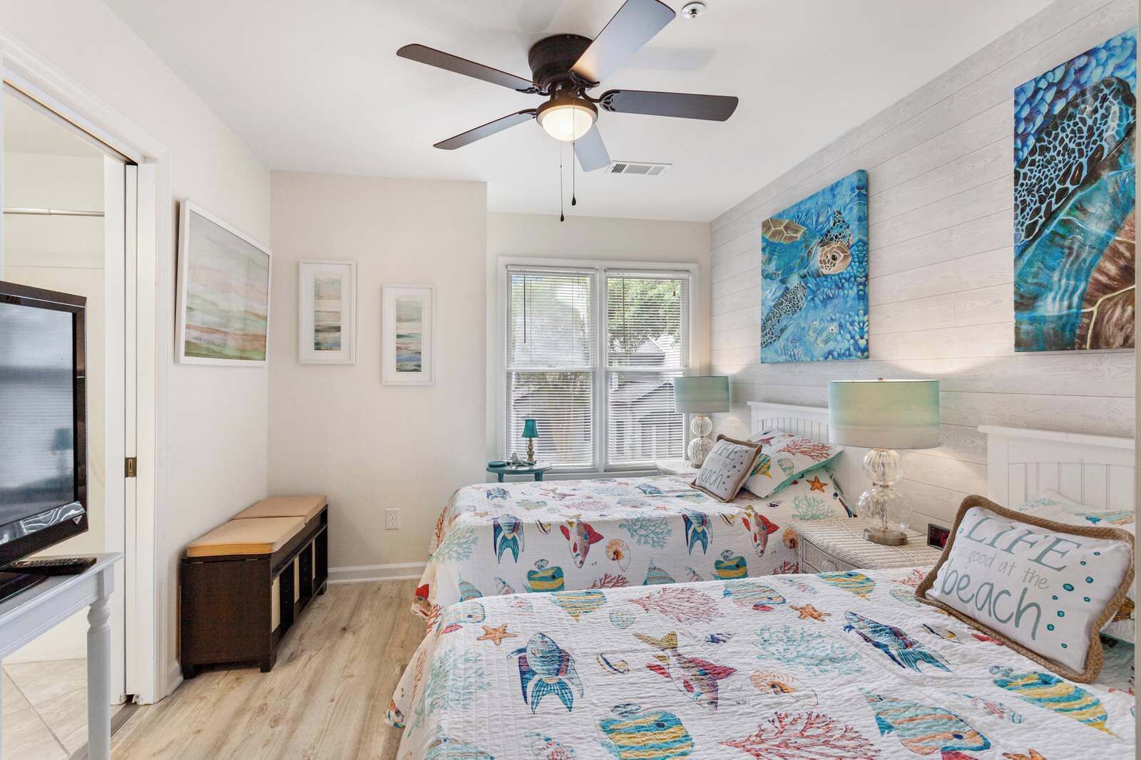 A bedroom with two beds, a ceiling fan, a TV, and artwork on the walls.