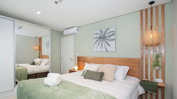 Ein modernes Schlafzimmer mit einem großen Bett, einem Holz-Kopfteil, einer Wand-Klimaanlage, einer Nachttischlampe und einer gerahmten Palmen-Wandkunst.