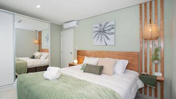 Ein modernes Schlafzimmer mit einem großen Bett, einem Holz-Kopfteil, einer Wand-Klimaanlage, einer Nachttischlampe und einer gerahmten Palmen-Wandkunst.