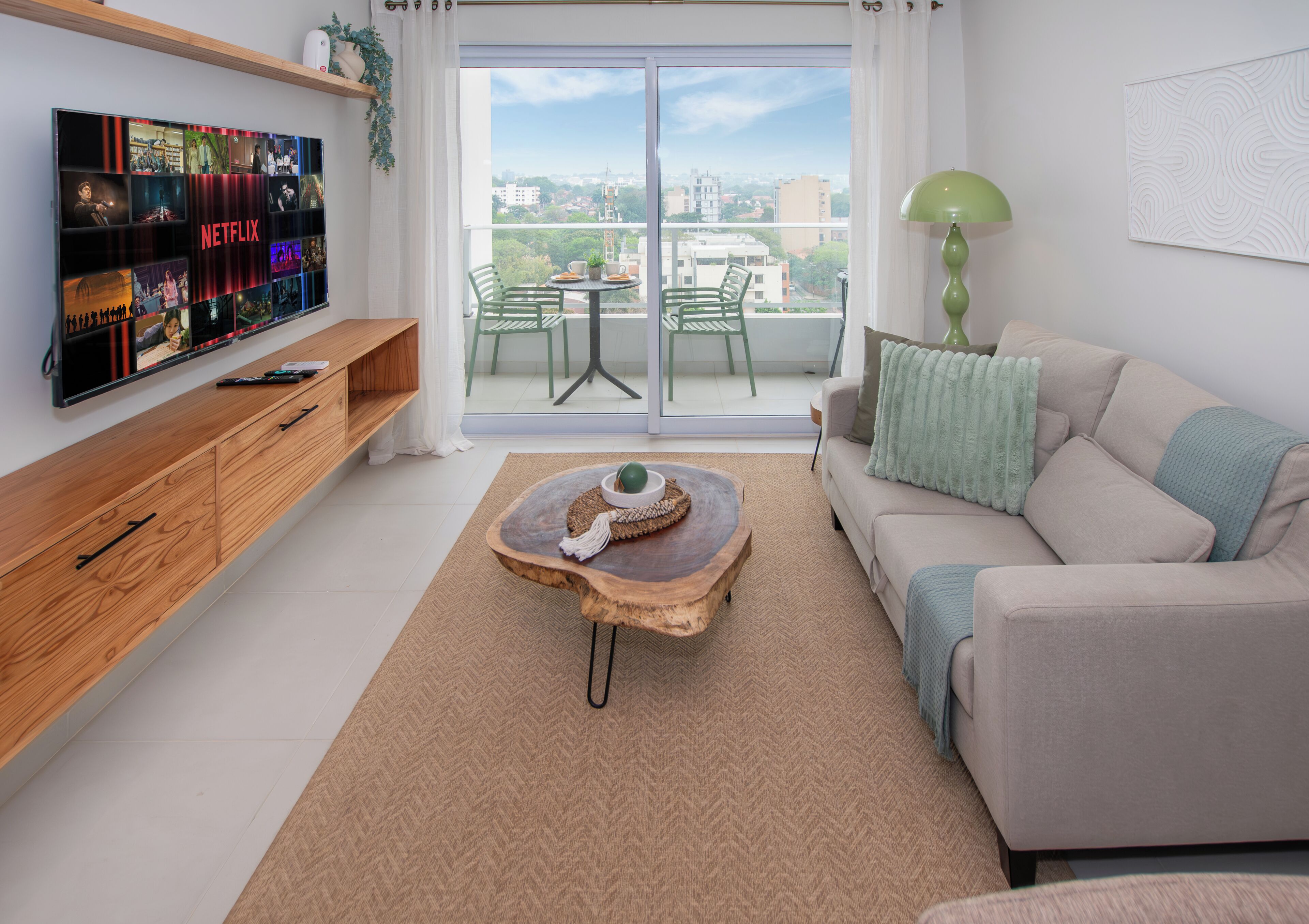 Ein modernes Wohnzimmer mit Fernseher, der Netflix-Inhalte zeigt, einem grünen Sofa, einem Holztisch und Blick auf die Stadt durch die Balkontüren.