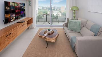 Ein modernes Wohnzimmer mit Fernseher, der Netflix-Inhalte zeigt, einem grünen Sofa, einem Holztisch und Blick auf die Stadt durch die Balkontüren.