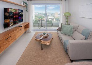 Un moderno salón con una televisión mostrando contenido de Netflix, un sofá verde, una mesa de centro de madera y vistas de la ciudad a través de las puertas del balcón.