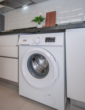 A front-loading white washing machine with a digital display, set in a kitchen with white cabinets and a countertop. - Corporate Stays First Living - 907 (Asunción)