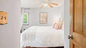 Una camera da letto con un letto, una ventola a soffitto, una finestra con persiane, un interruttore della luce fissato al muro e una porta.