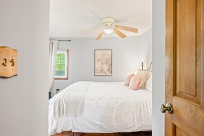 Ein Schlafzimmer mit einem Bett, einem Deckenventilator, einem Fenster mit Jalousien, einem an der Wand montierten Lichtschalter und einer Tür.
