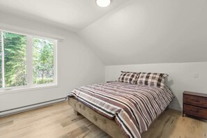 Et soveværelse med skråvæg, stort vindue, seng med stribet sengetøj og et natbord.