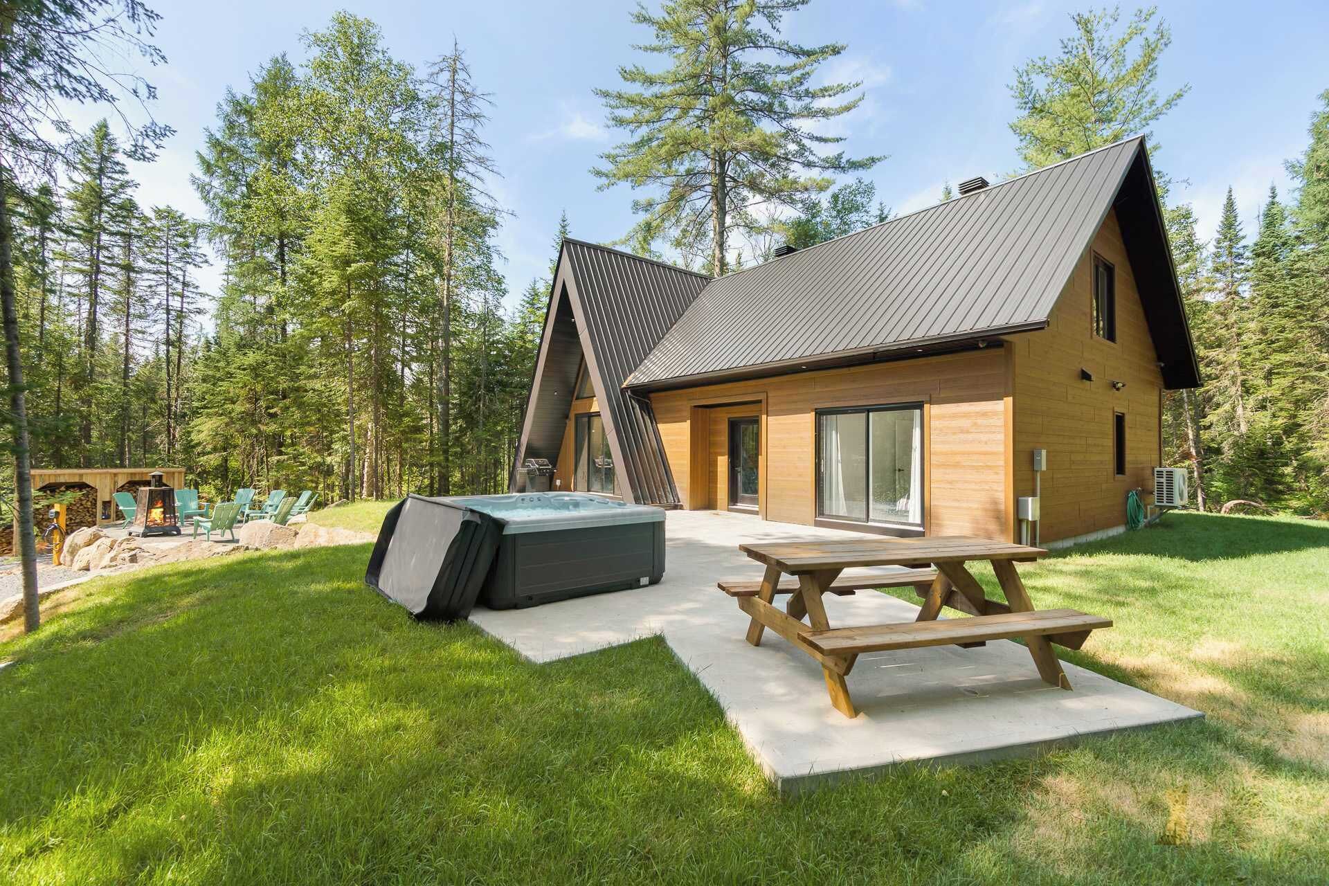 A modern cabin with a sloped roof, a hot tub, and a picnic table in a forested area.