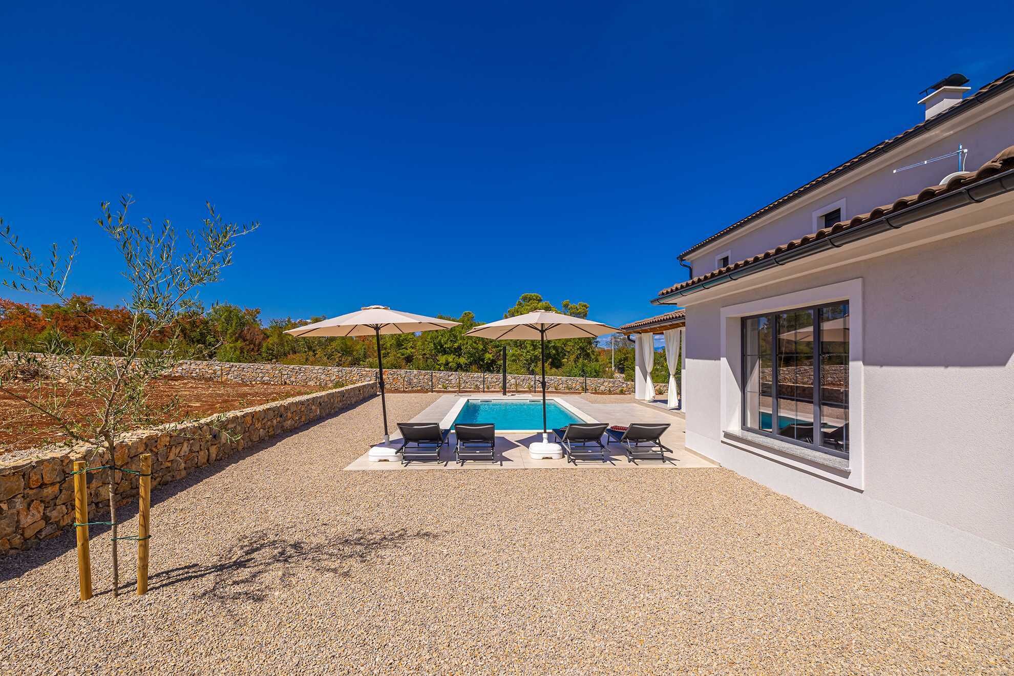 A pool area with lounge chairs and umbrellas, a stone wall, and a building with a tiled roof.