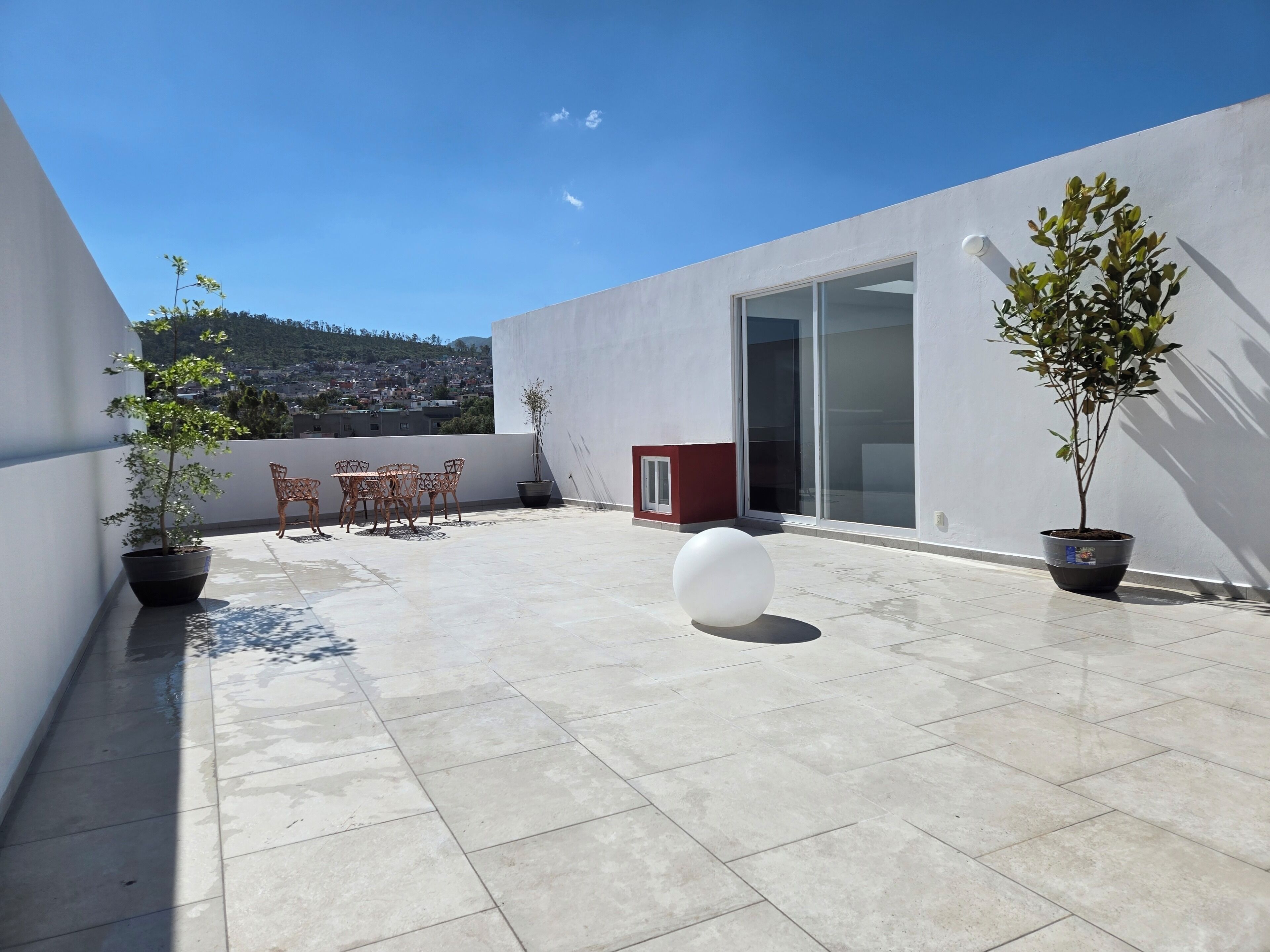 A rooftop terrace with a table and chairs, potted plants, and a view of a hillside town.