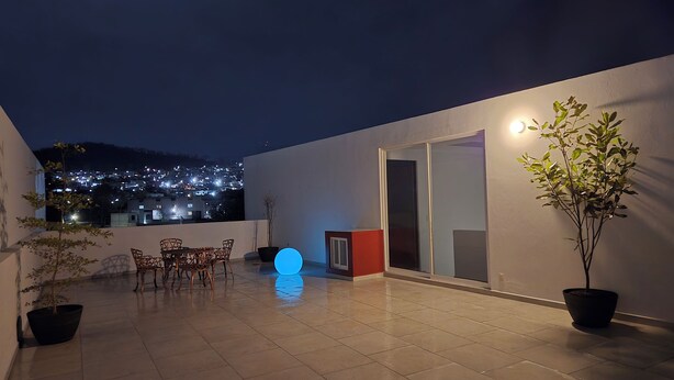 A rooftop patio with a table and chairs, a potted plant, and a view of a cityscape at night.
