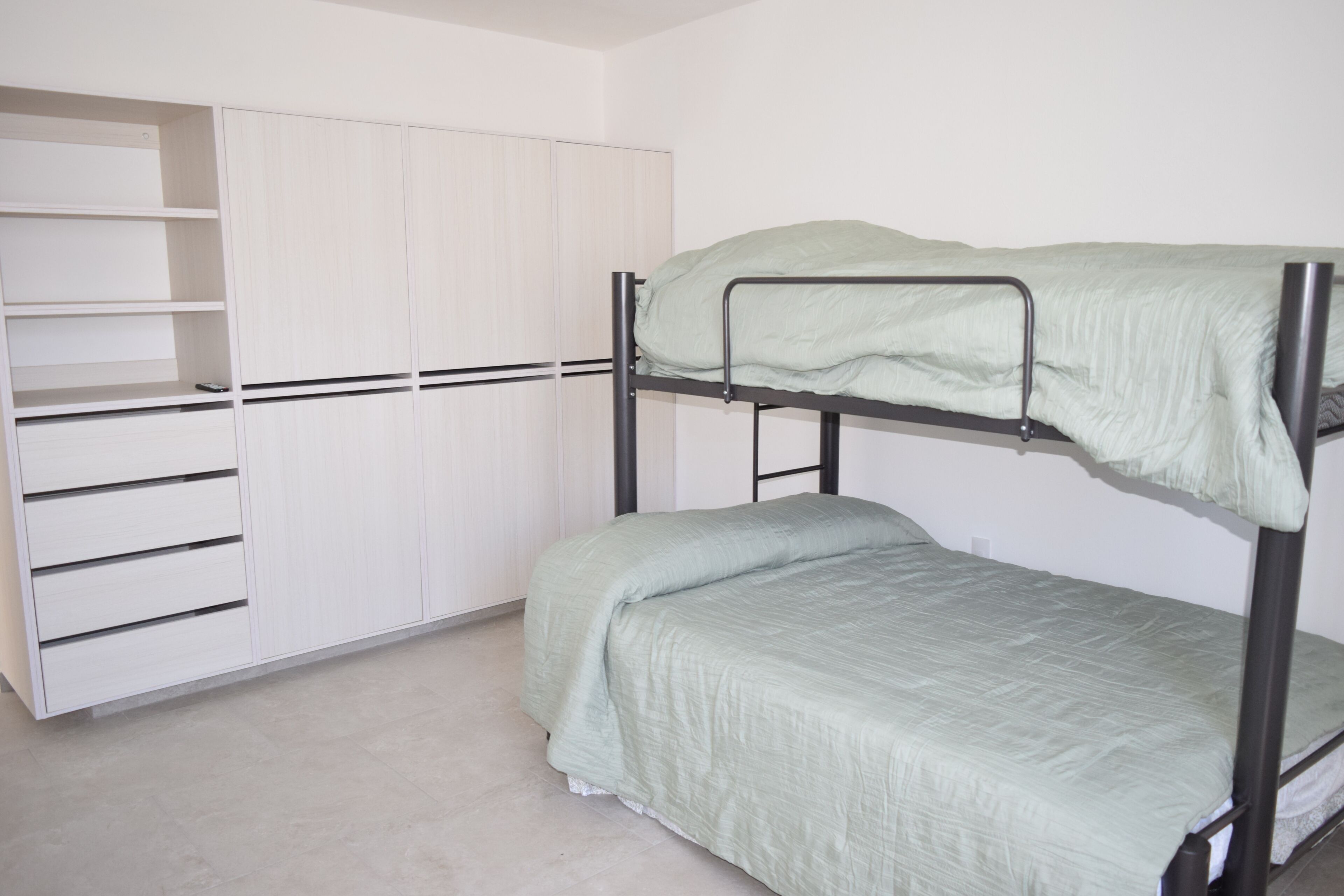 A bunk bed with a green mattress, a white wardrobe with multiple shelves, and a tiled floor.