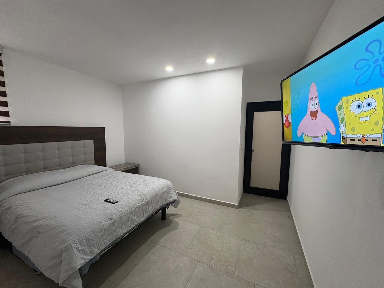 A bedroom with a bed, a nightstand, a TV mounted on the wall, and a door.