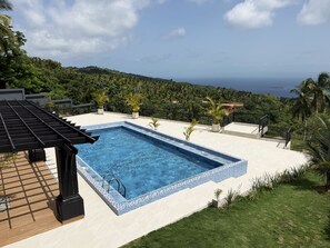 Ein rechteckiger Pool mit Fliesenrand, umgeben von einer weißen Liegefläche, mit Blick auf den Ozean und Hügel im Hintergrund.