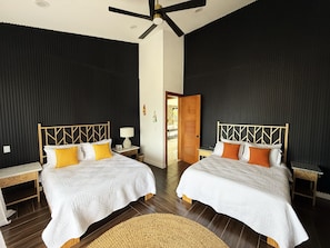 Een hotelkamer met twee bedden, een plafondventilator, een houten deur en een klein tafeltje met een lamp.