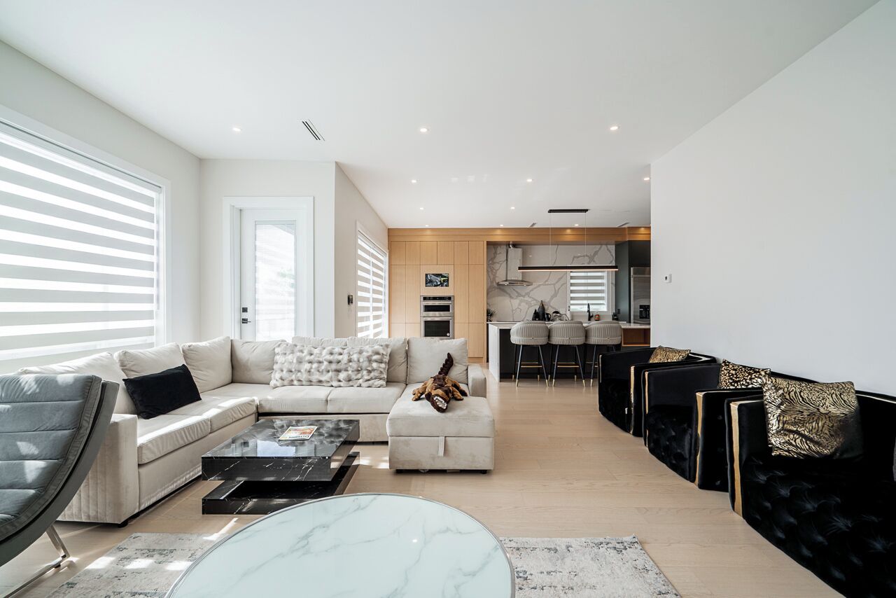 A modern living room with a sofa, ottoman, and coffee table, a kitchen in the background, and a dining area with chairs.