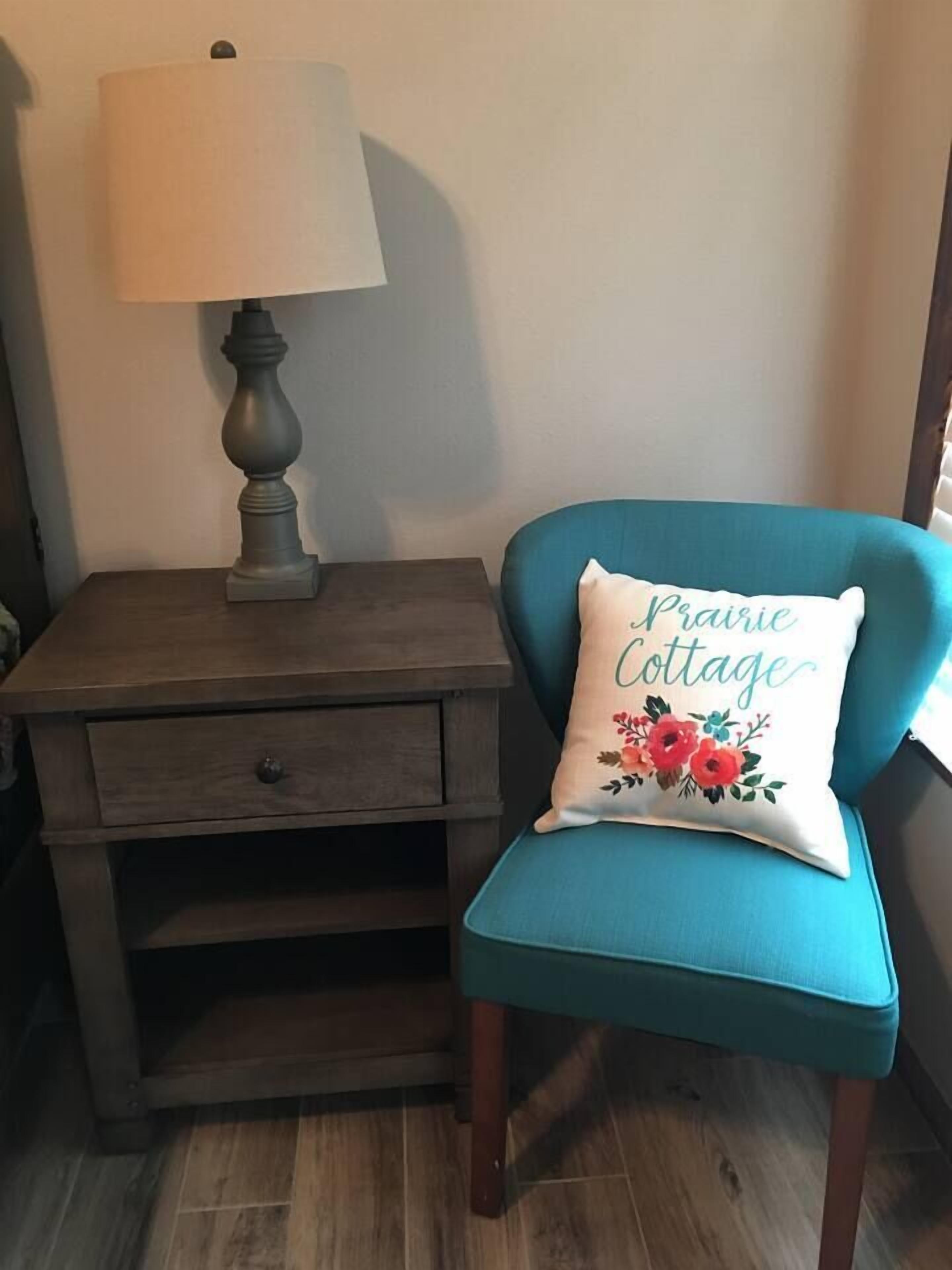 A teal armchair with a decorative pillow, a wooden nightstand with a lamp, and a drawer.