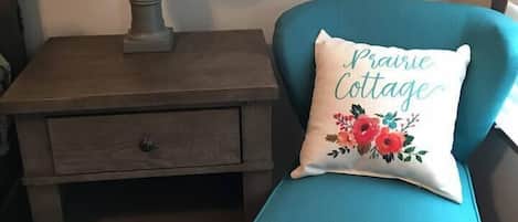 A teal armchair with a decorative pillow, a wooden nightstand with a lamp, and a drawer.