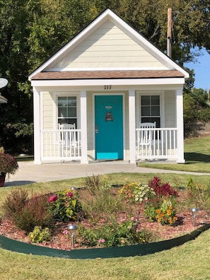 A small, single-story building with a white exterior and a teal door, surrounded by a landscaped garden with various plants and flowers. - Prairie Cottage King Suite 1/2 block from PW Merc (Pawhuska)