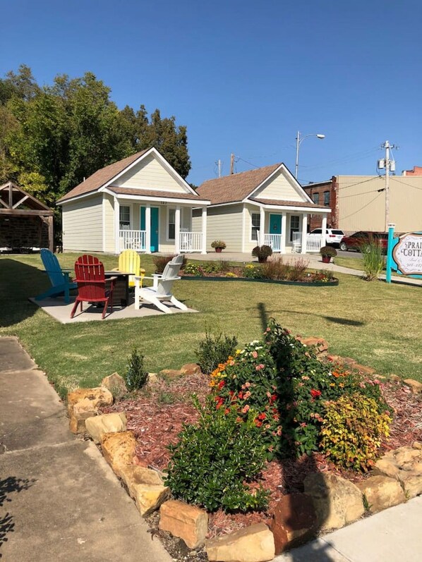 A two-story cottage with a porch, a lawn with colorful Adirondack chairs, and a flower bed.