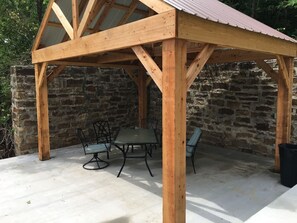 A wooden gazebo with a stone wall backdrop, a table with chairs, and a trash bin.